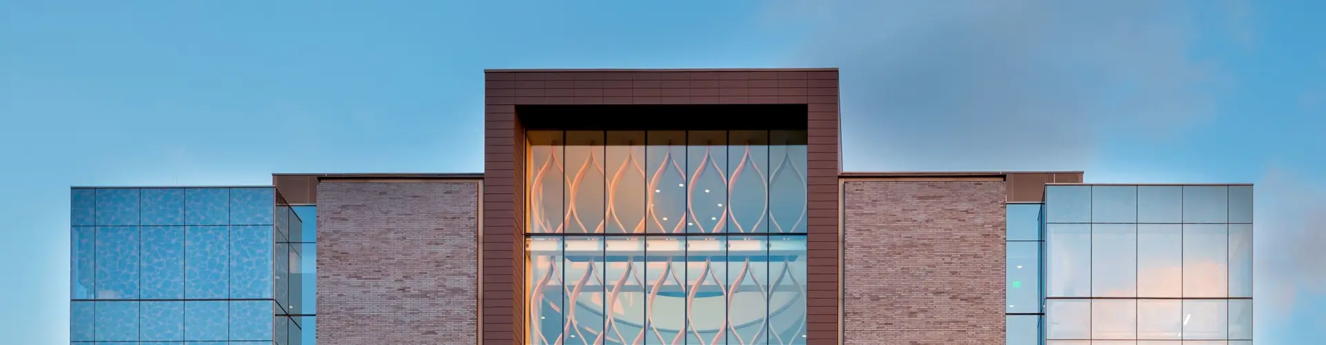 The image shows the modern facade of a building with brick walls, large glass windows, and a central section featuring patterned glass panels against a blue sky.
