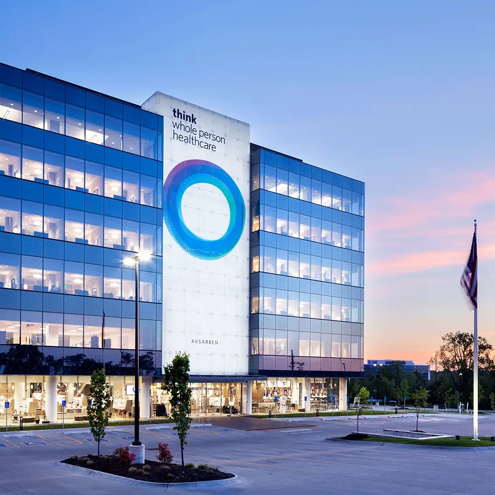 A modern glass office building at sunset displays the words think healthcare and a large blue circular design on its facade. An American flag stands in the foreground near an empty parking lot.