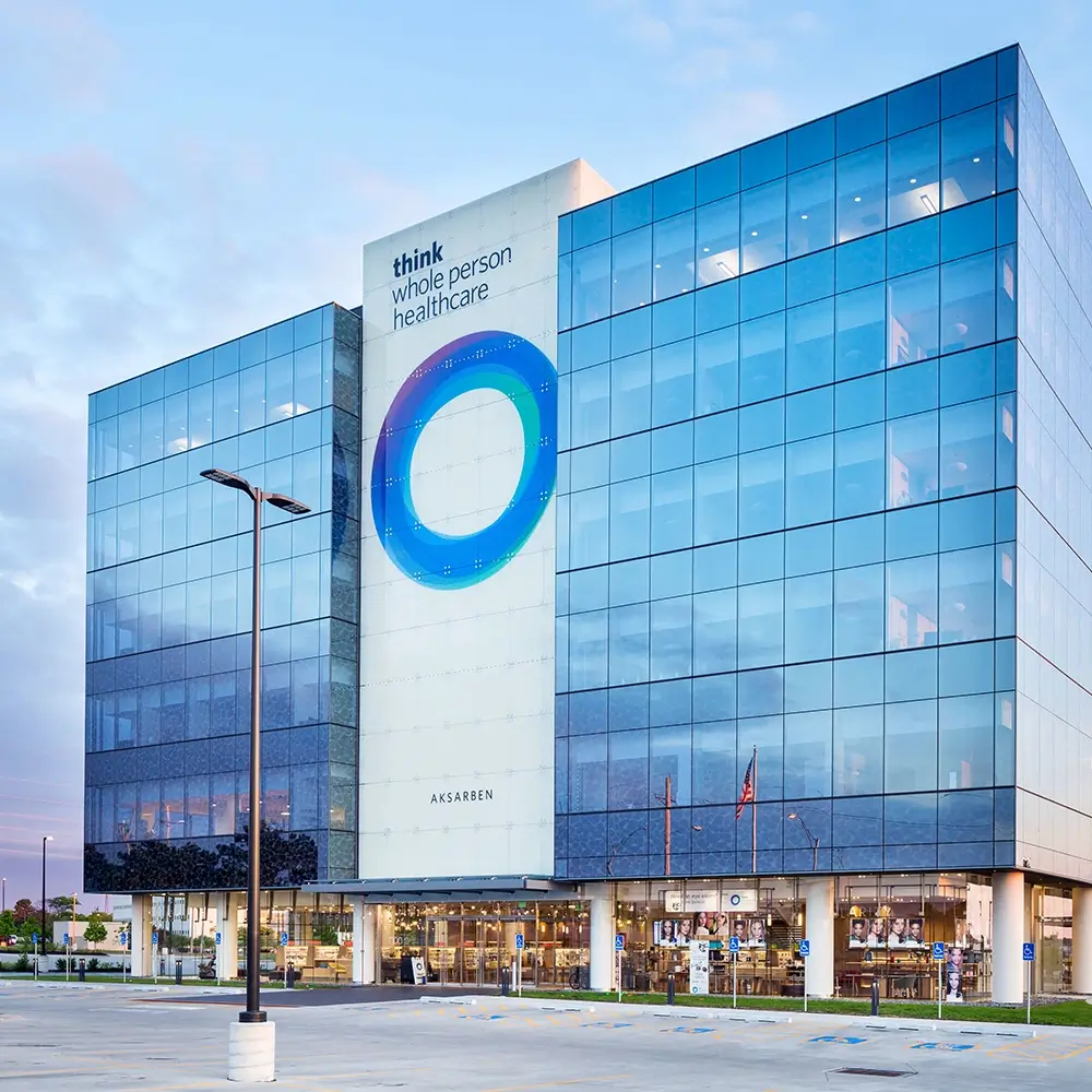 A modern glass office building with a large circular blue logo and the words think healthcare on the facade. The building features multiple stories, expansive windows, and a parking lot in front.